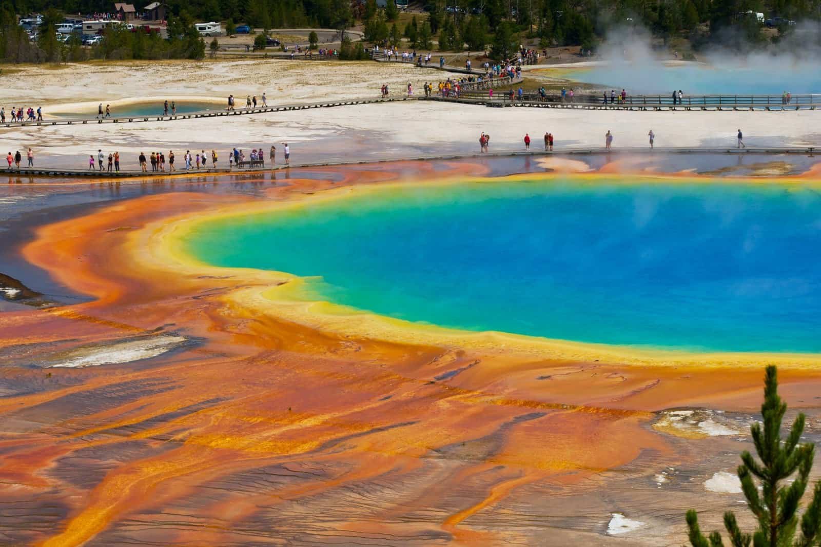 Visitors walk along boardwalks beside the vibrant Grand Prismatic Spring, showcasing vivid rainbow colors and steaming geothermal pools—one of the most breathtaking vacation sites in the USA.